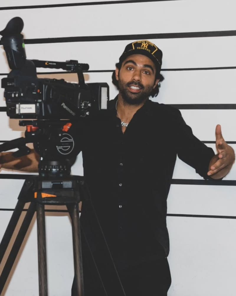 Shawn Regruto in a Yankees cap, arms wide, standing behind a video camera on a tripod in front of a height chart.