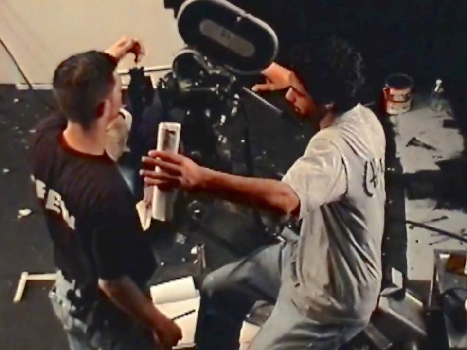 Shawn Regruto on a film set, leaning into a large film camera beside a crew member.